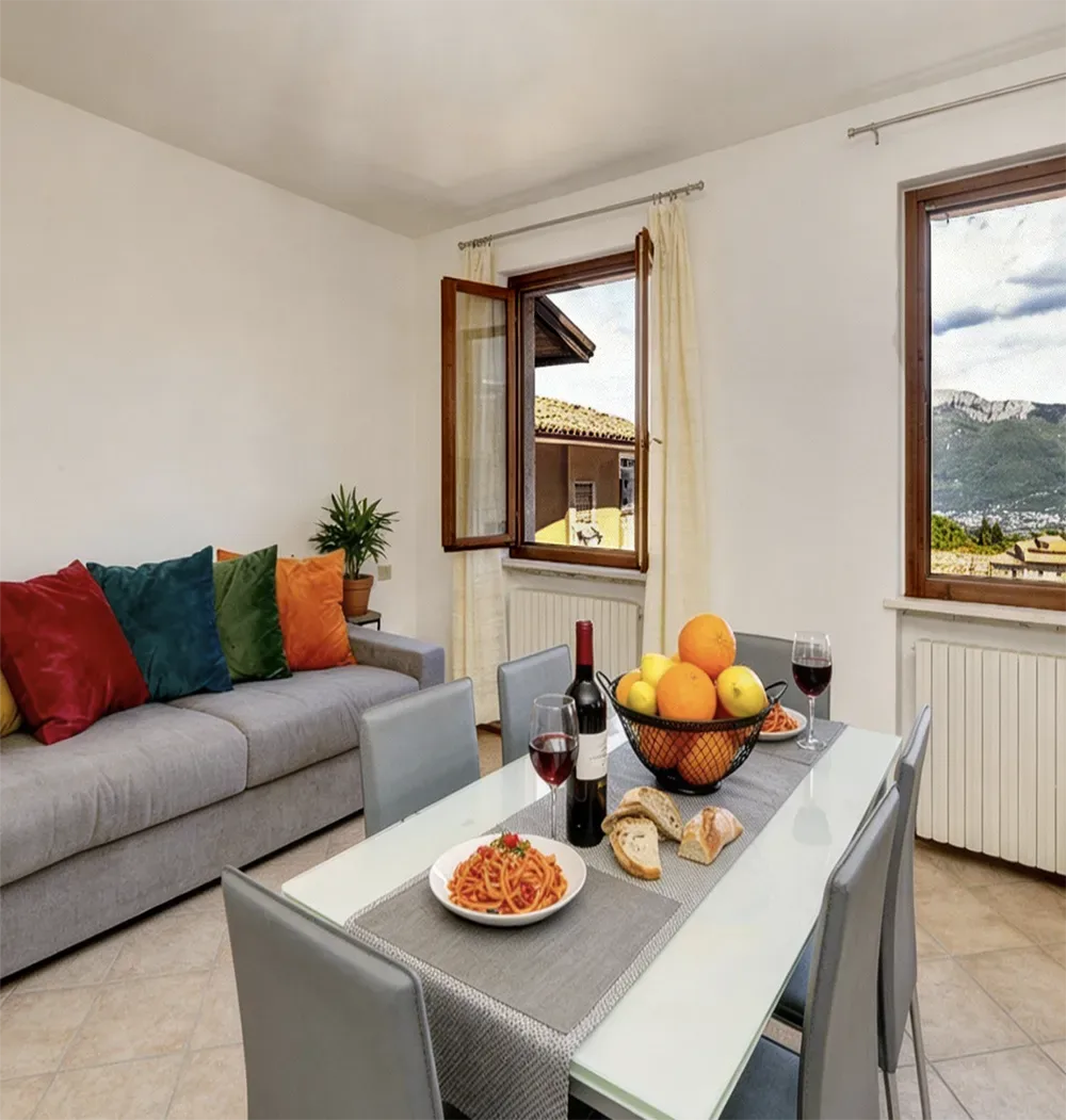Bright dining area with pasta, wine and bread, window view of Lake Garda and mountains